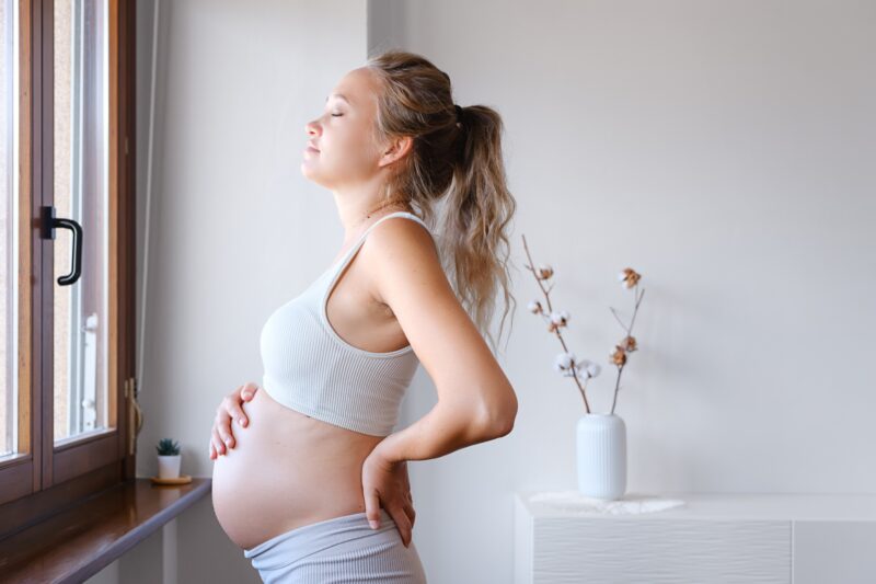 A pregnant woman stands looking out of the window, with her hand resting on her stomach.