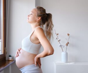 A pregnant woman stands looking out of the window, with her hand resting on her stomach.