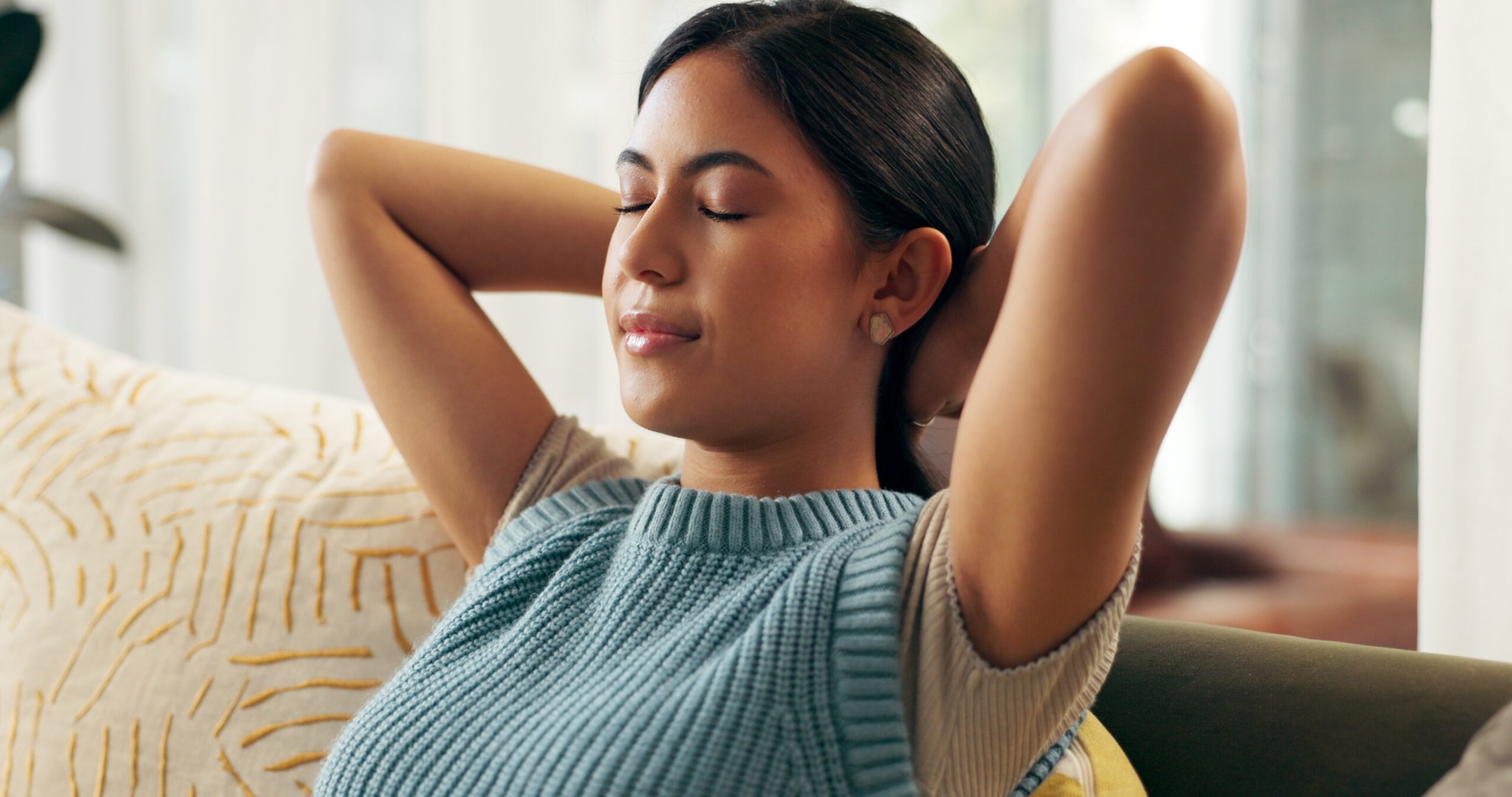 A woman sits meditating with her eyes closed and arms behind her head.
