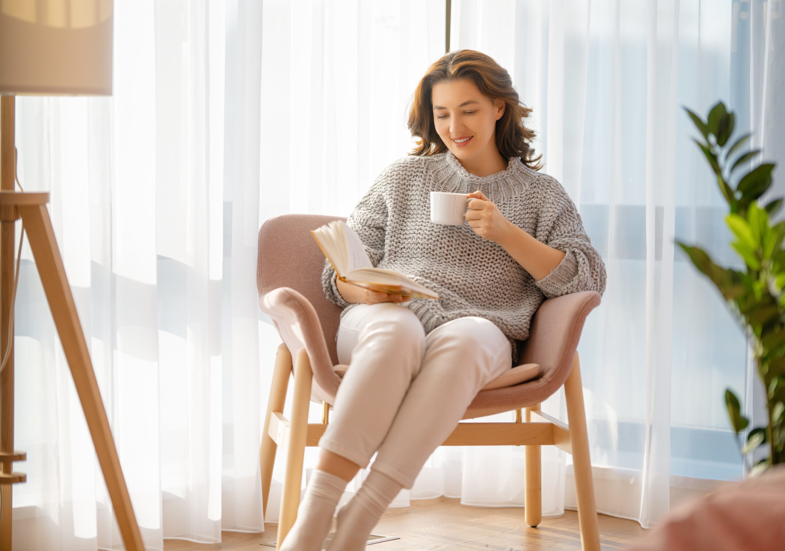 A woman sits reading a book and drinking a cup of adaptogenic tea in a calm, minimal room.