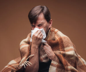 A man is wrapped in a blanket blowing his nose. self-care for cold and flu season