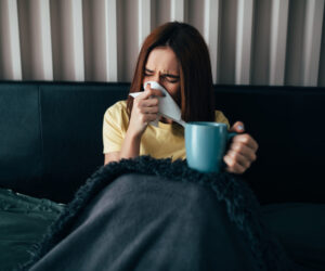 An unwell woman sits in bed blowing her nose and drinking a hot cup of herbal tea for hydration and immunity