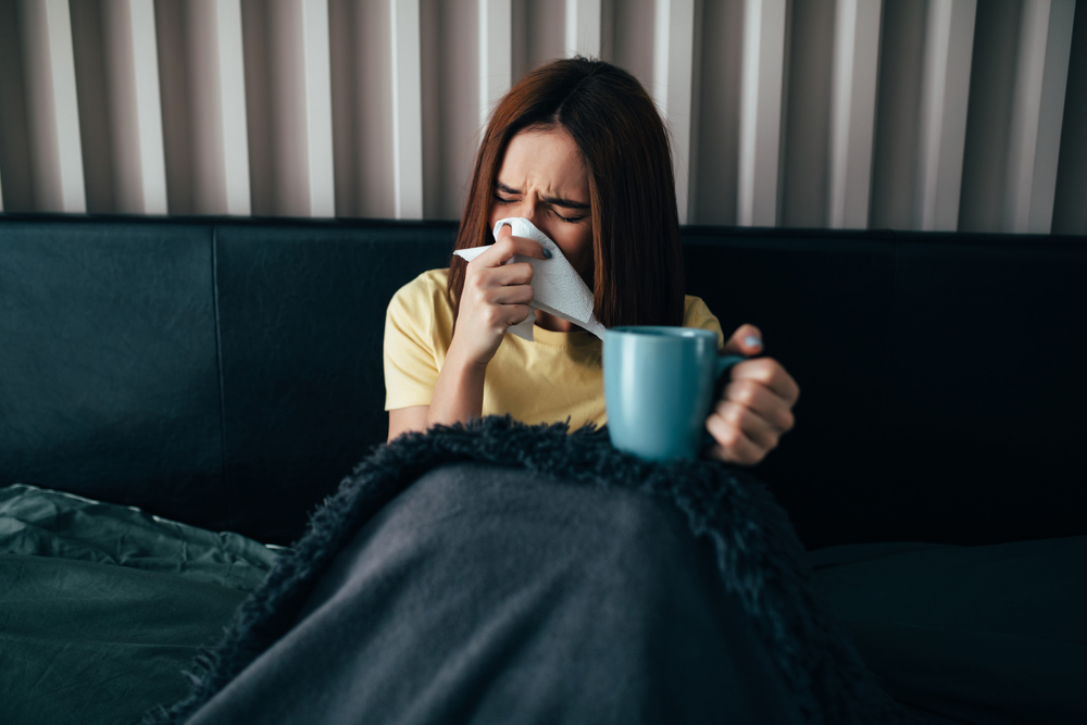 An unwell woman sits in bed blowing her nose and drinking a hot cup of herbal tea for hydration and immunity